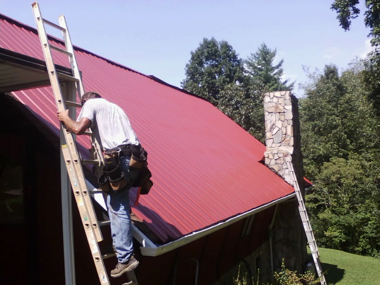 Roofer on a ladder beside a red metal roof and stone chimney — Soffit & Fascia Repair by Roofing Brothers in Churchville